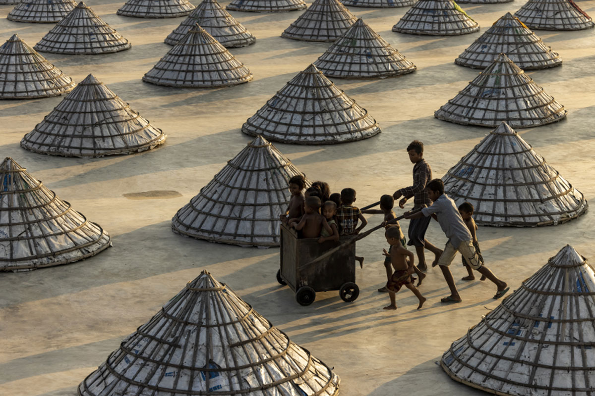 Childhood, capturing children weaving in and out of scores of giant cones (topas) in a Bangladeshi rice processing plant as they play with a cart used by workers to carry and dry rice. © Azim Khan Ronnie, Bangladesh, Winner, Open Competition, Lifestyle, Sony World Photography Awards 2023