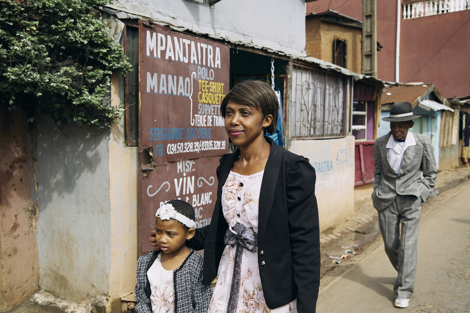 Outside a building with signs and vegetation, a woman in a floral dress and a child in a matching outfit walk by. The woman looks ahead confidently while the child seems pensive. In the background, the elderly man from the first photo walks away, creating a narrative connection between the images.