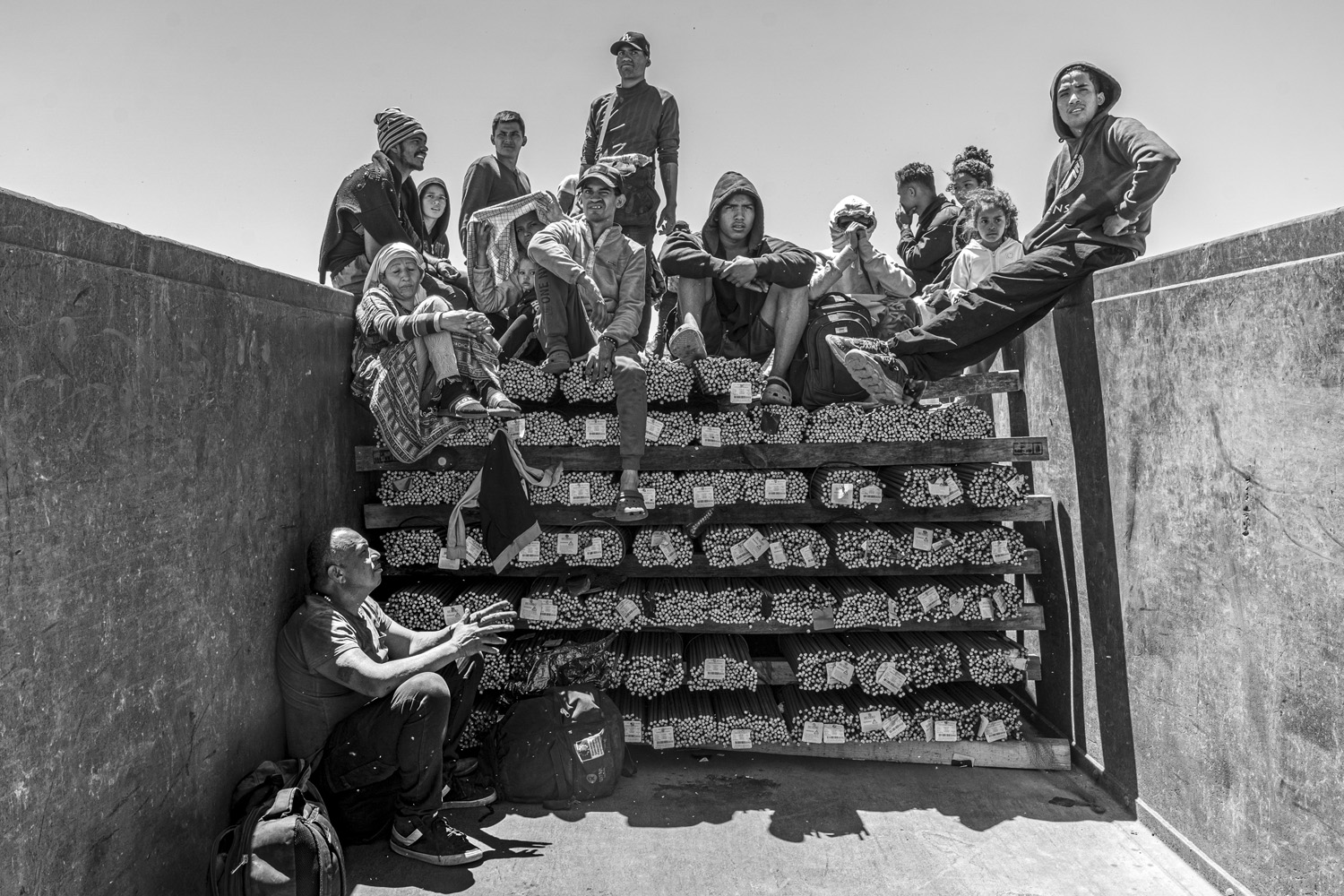 A black and white photo of migrants seated in a tiered fashion on a truck full of goods. Various expressions of fatigue and concern are visible among the group