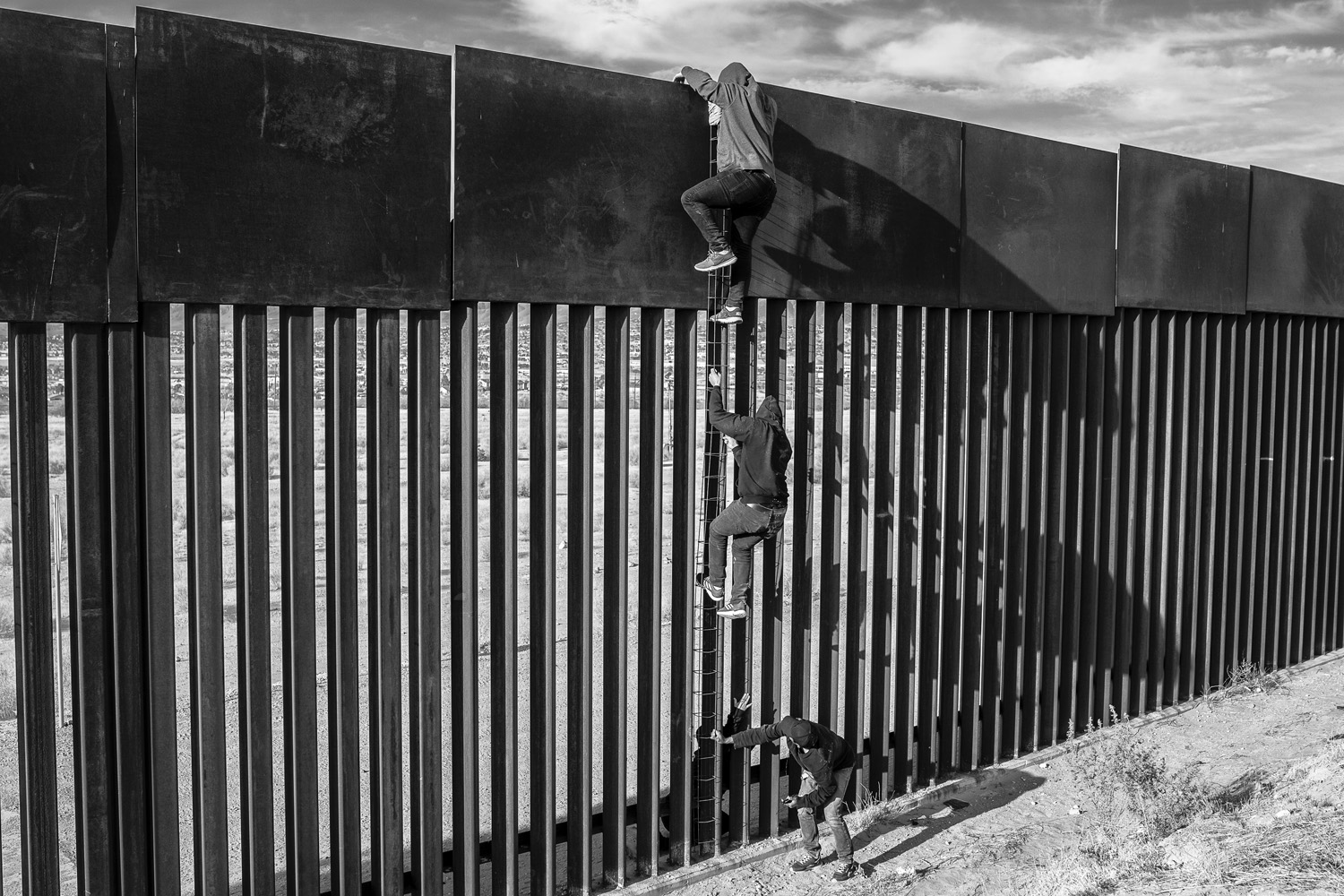 Two individuals are seen climbing a tall, vertical metal barrier using a rudimentary ladder. The stark black and white contrast highlights the effort and determination involved in overcoming physical and metaphorical boundaries.