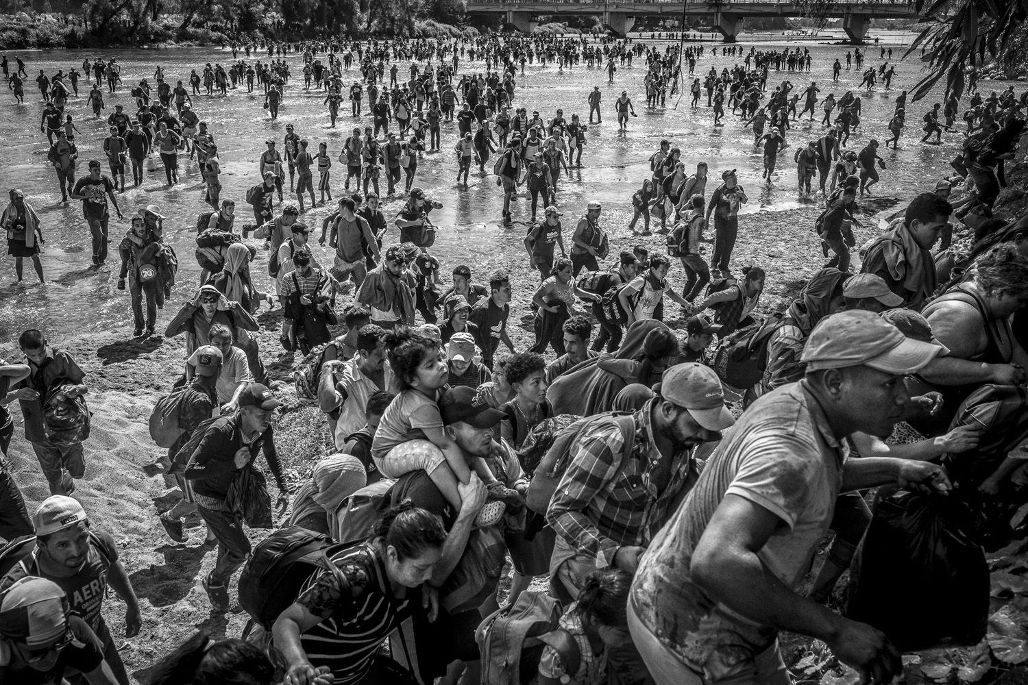 A crowded scene in black and white, depicting numerous people wading through a shallow river. The chaos and urgency of movement are palpable as individuals carry belongings and help one another navigate the waters, illustrating the intensity and scale of human migration.