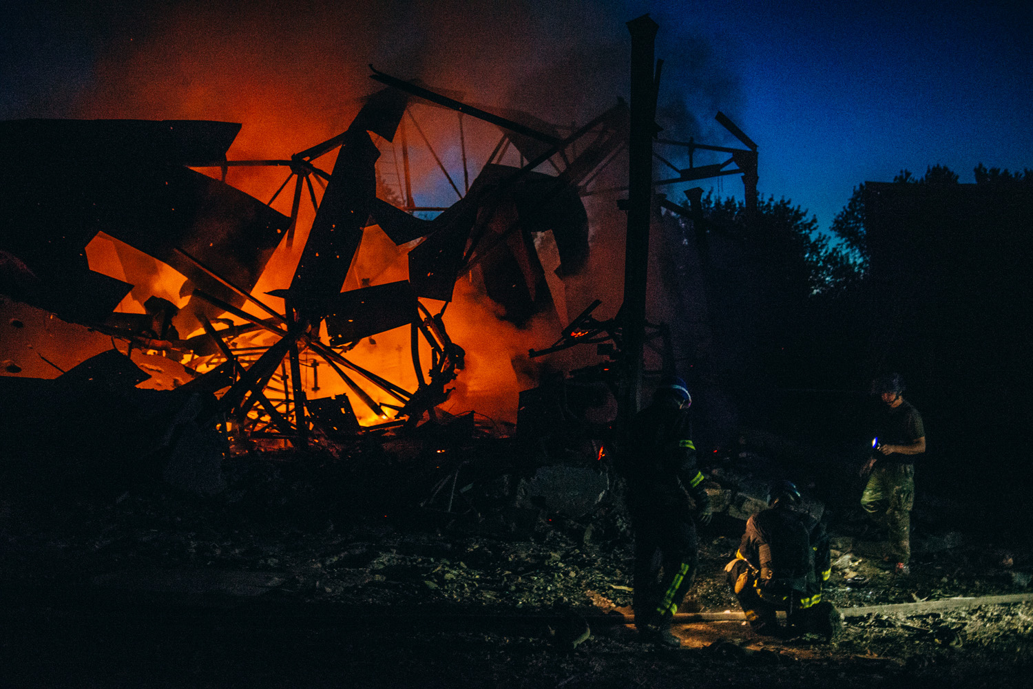 In the image, firefighters are seen at night battling a fierce blaze that engulfs what appears to be a collapsed building in a war zone. The flames illuminate the scene with an intense orange glow against the night sky, while silhouettes of the firefighters are depicted working amidst the destruction, highlighting their courage in the face of danger.