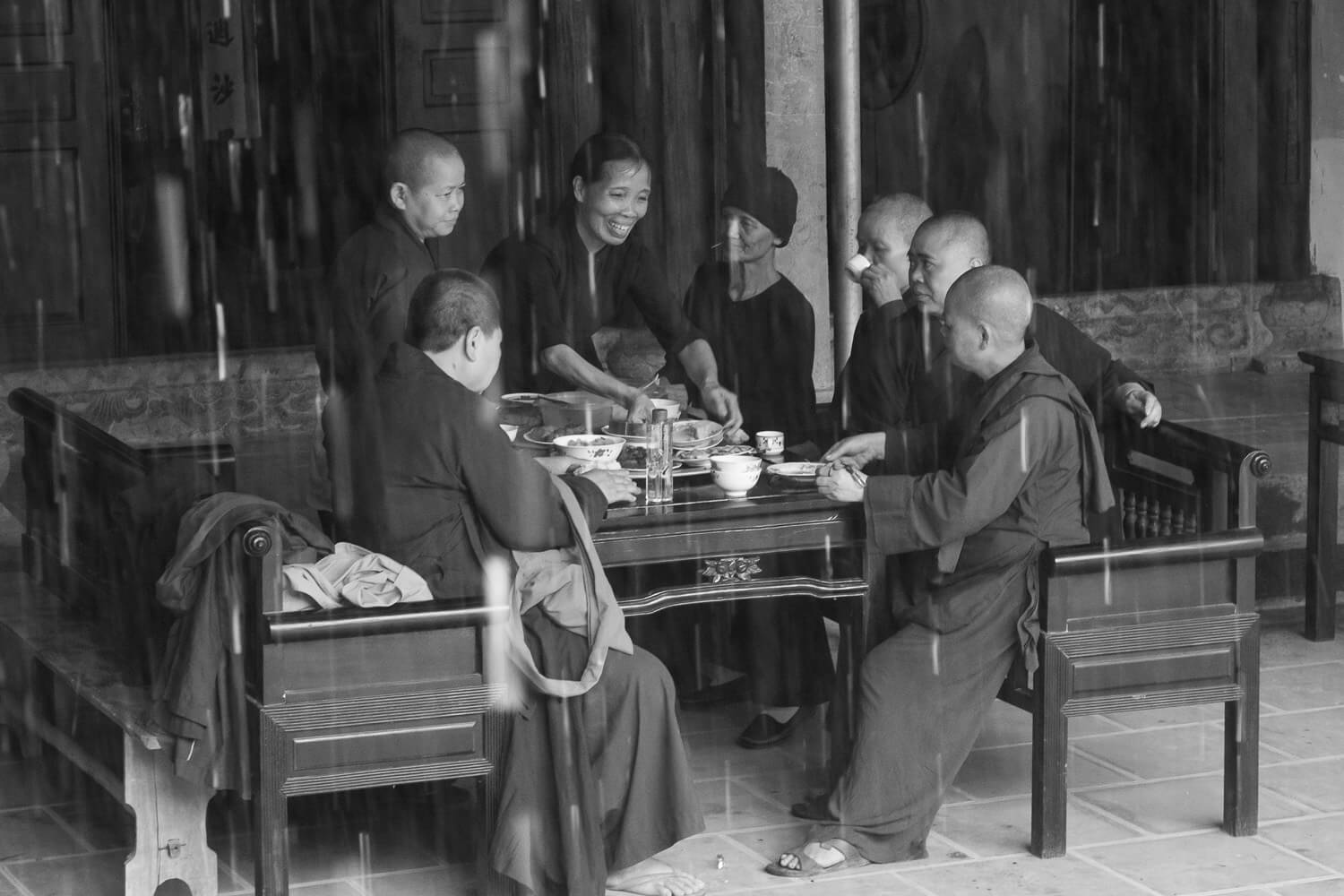 A group of Buddhist monks in traditional attire is seated around a wooden table, sharing a meal in a serene, open-air setting that appears to be within a temple's confines, as seen through the grayscale filter, adding a timeless quality to the moment.