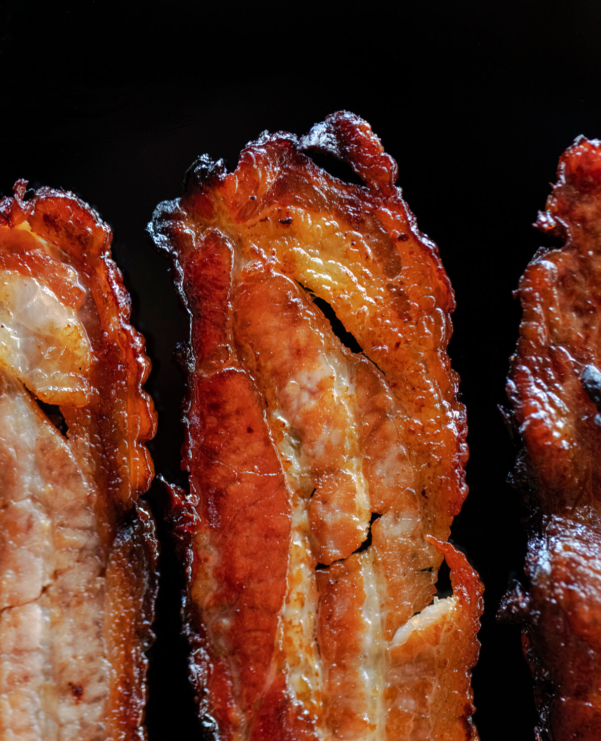 Crispy slices of bacon are showcased in close-up against a dark background, highlighting the textures and glossy sheen of the cooked meat with a play of light and shadow enhancing its savoury appeal.