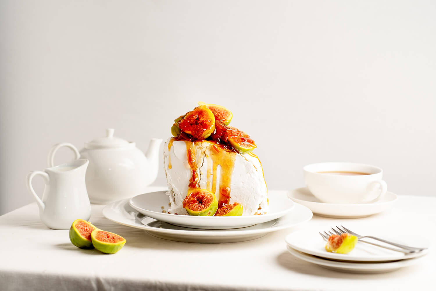 A pavlova cake with crisp white meringue is topped with fresh fig slices and a drizzling of honey, presented on a white plate. The table setting includes a teapot, milk jug, teacup, and extra plates, all in clean white, with a neutral background accentuating the dessert’s vibrant colors and textures.