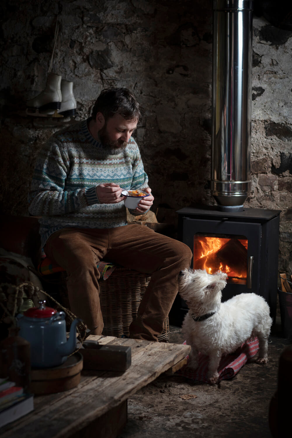 A man in a patterned sweater sits comfortably on a wicker chair, eating from a bowl in a rustic stone cottage, with a wood-burning stove and a small white dog looking up at him, creating a cozy and intimate atmosphere.