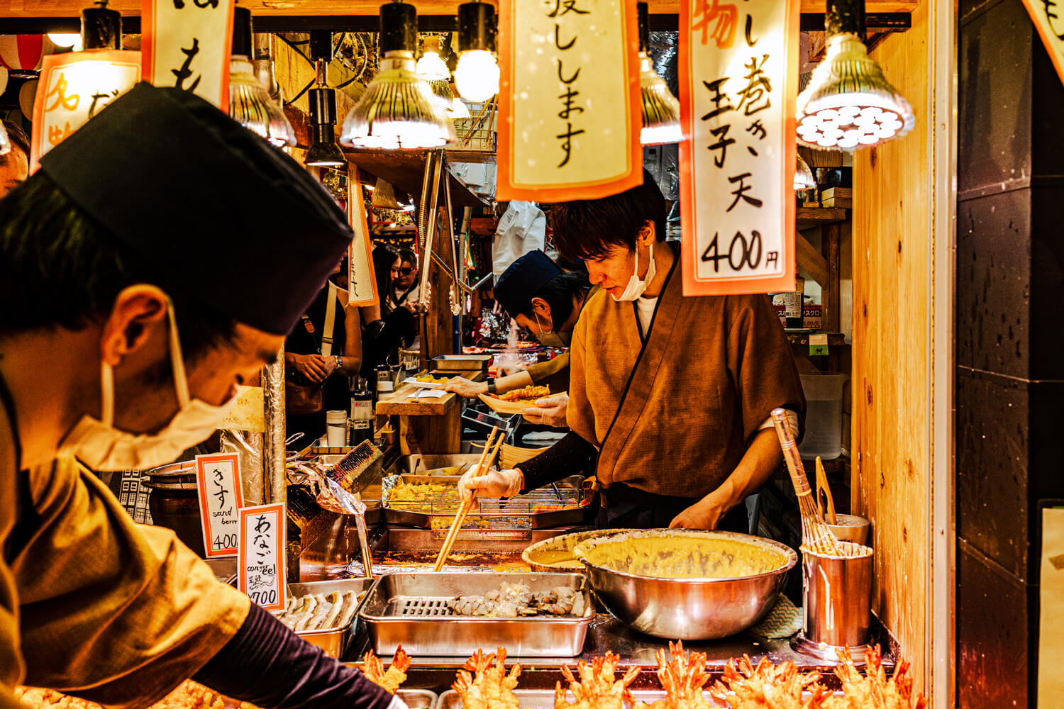 Bustling with activity, a Japanese street food stall glows warmly under hanging lamps, with chefs in traditional attire busily preparing food. Lanterns with Japanese script hang above, and an array of seafood is on display, inviting passersby to indulge in the local cuisine.