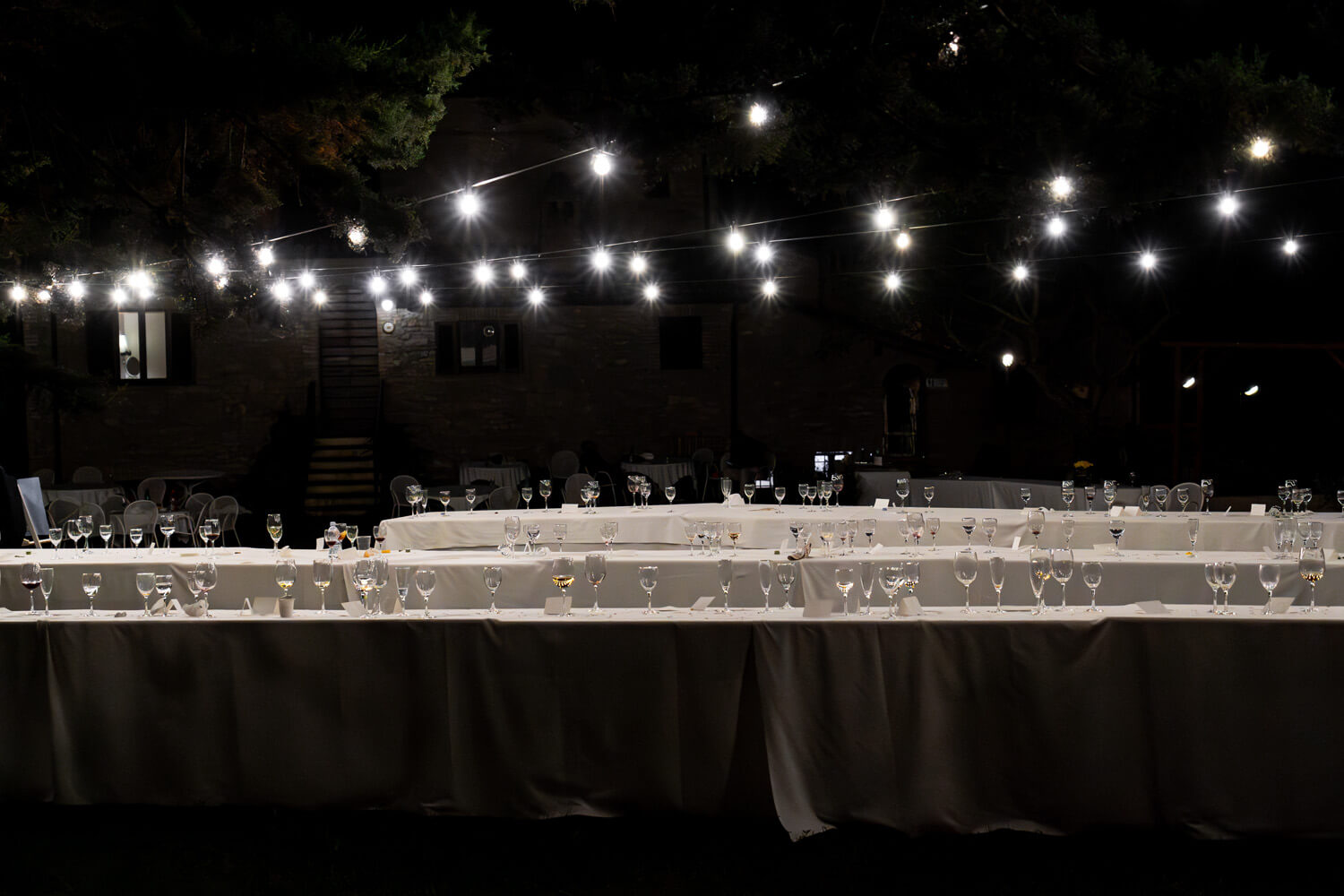 An elegantly set long dining table awaits guests at an outdoor evening event, with strings of lights creating a festive canopy above. The soft glow against the night sky lends an intimate ambiance to the setting.