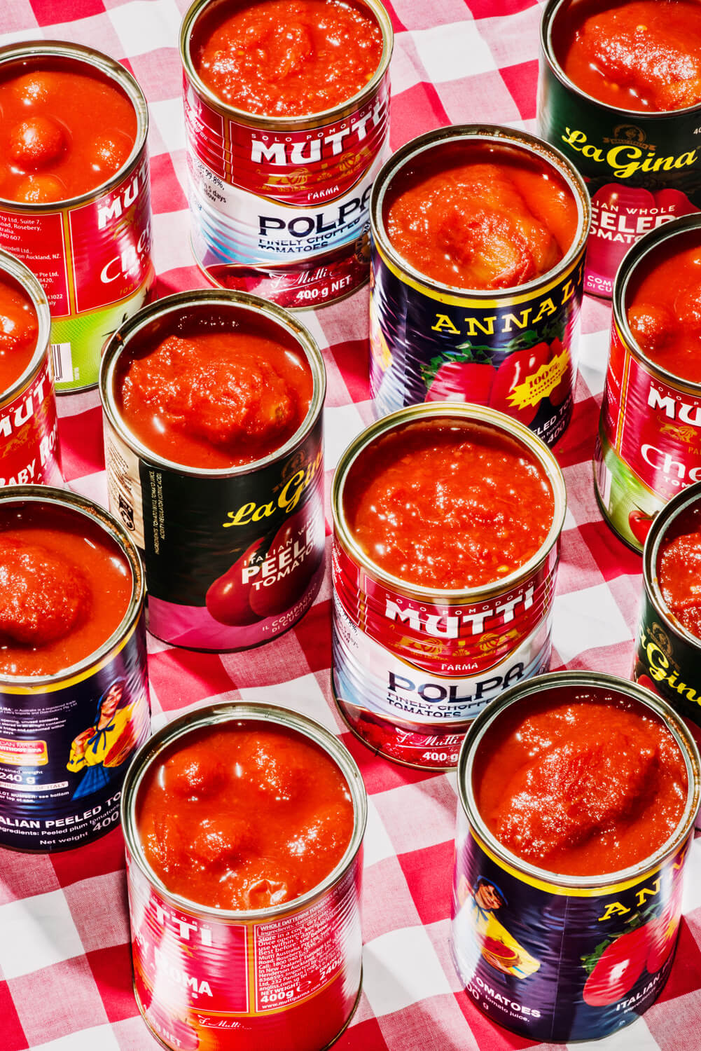 Various cans of tomato products, with vibrant labels, are open and arranged on a classic red and white chequered tablecloth, the rich red of the tomatoes standing out against the pattern, suggestive of preparation for a flavourful cooking session.