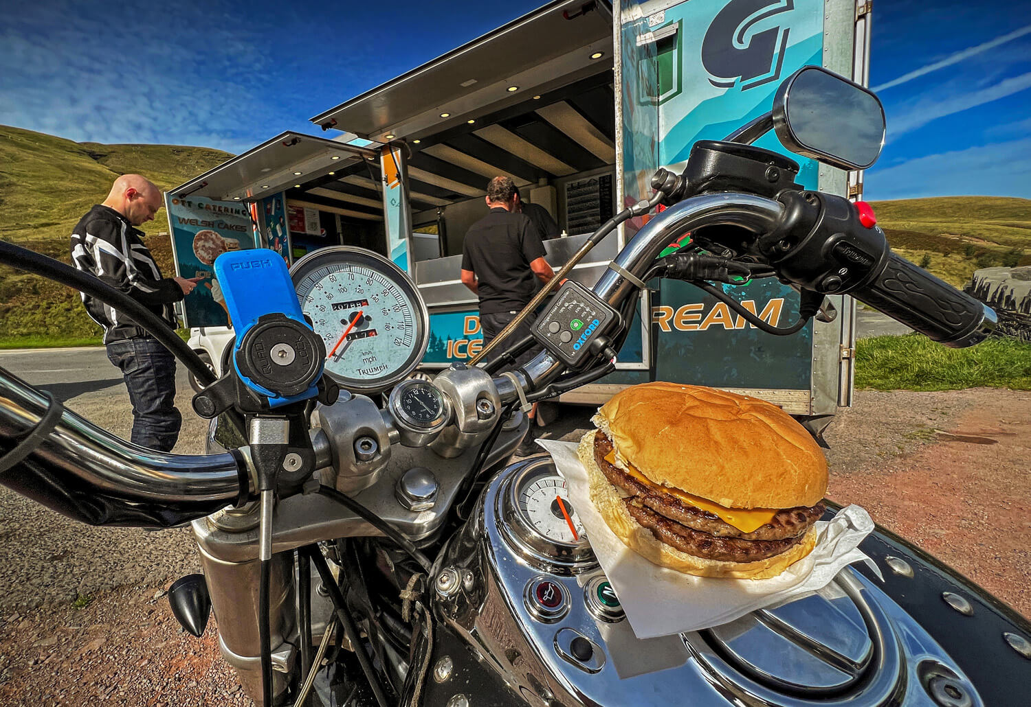 From the rider's perspective, the image showcases a tantalizing cheeseburger resting on a motorcycle tank in clear weather. The bike's chrome details gleam under the sunlight, and a food truck and fellow biker are visible in the background, suggesting a roadside stop during a journey.