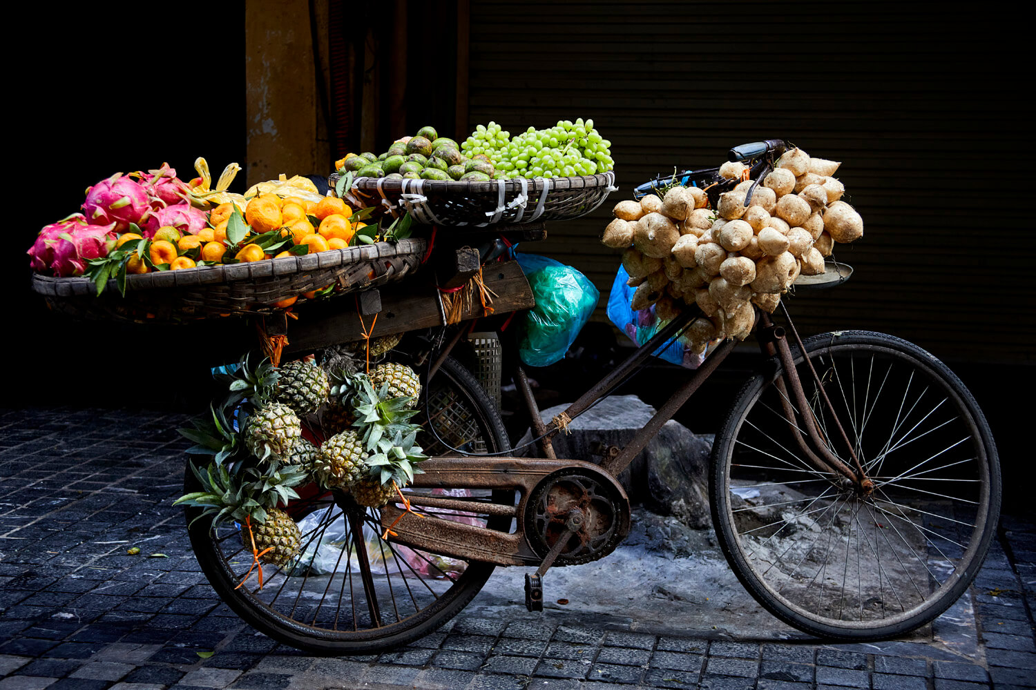 This image features a laden bicycle, standing as a makeshift market stall, against a dark background. The bike is heavily adorned with an assortment of colourful fruits: dragon fruits, bananas, grapes, and pineapples, with bunches of white bulbs hanging from the handlebar.