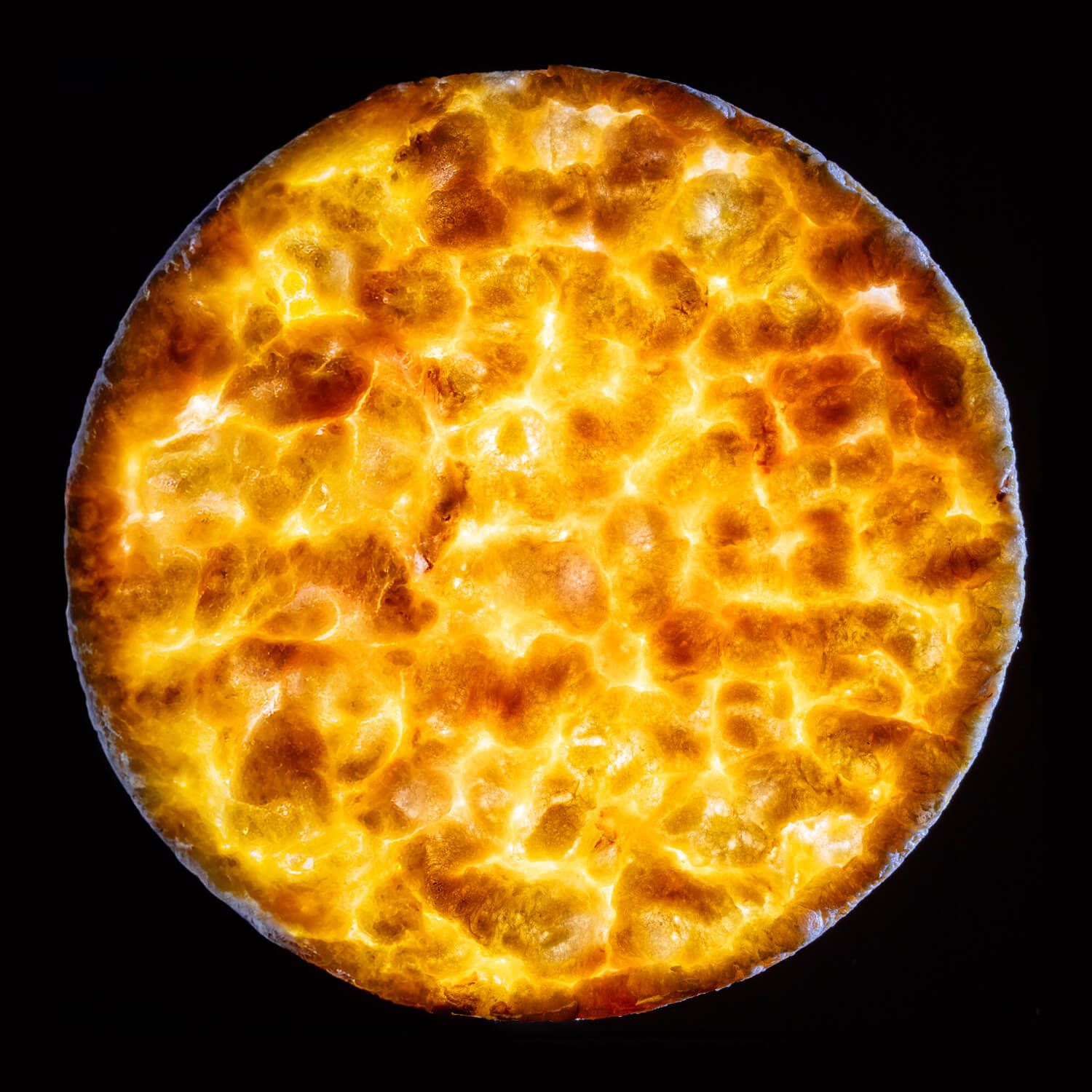 The image showcases a close-up of a round, golden rice cracker back lit against a dark background, highlighting the textures and vibrant colours of the crust. The surface appears bubbly and crisp, indicative of a flaky or cheesy top that's been browned to perfection, and also reminiscent for the surface of the moon.