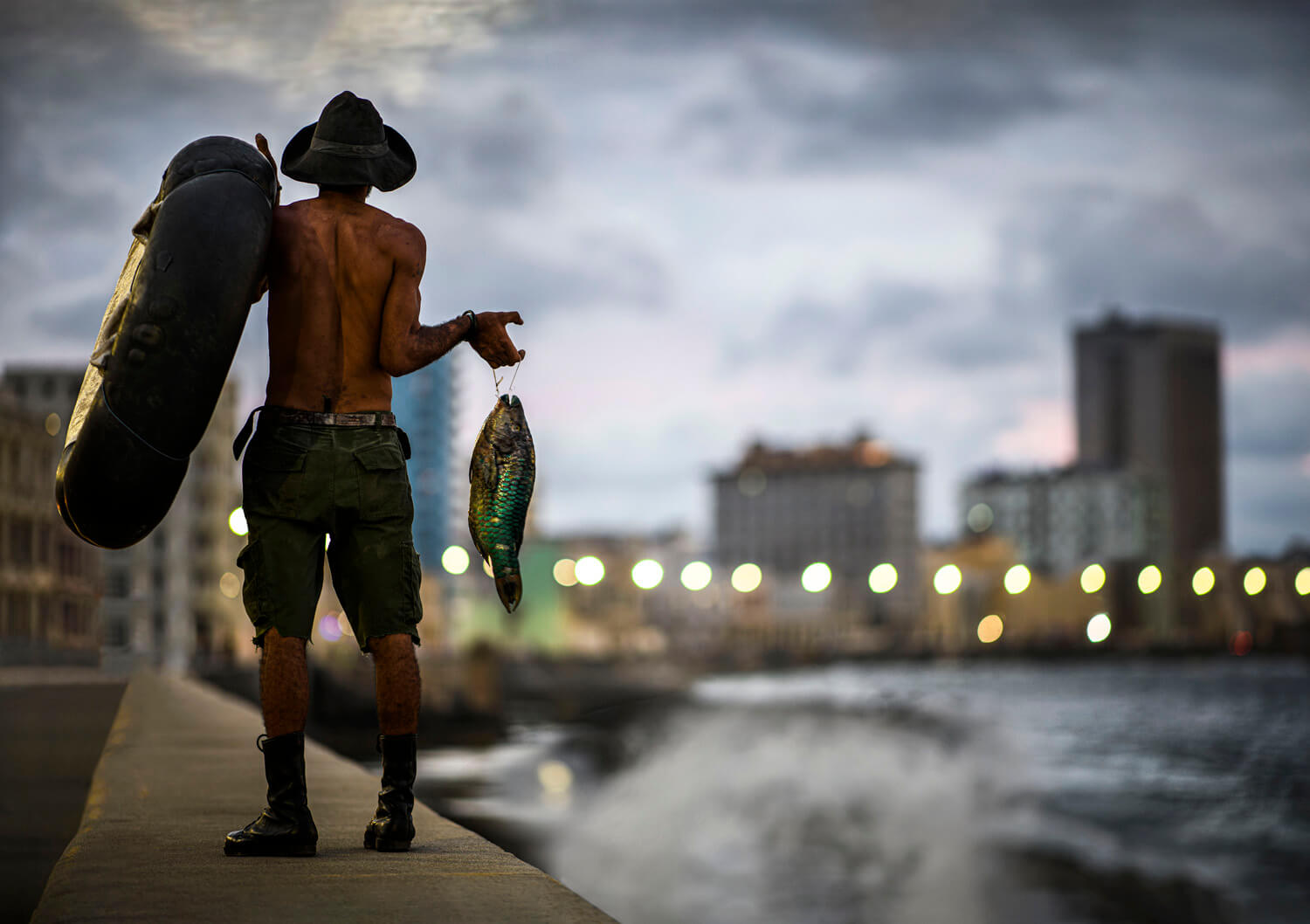 This image captures a man from behind as he walks along a waterfront promenade at dusk. He is shirtless, showcasing a toned physique, and wears a wide-brimmed hat and cargo shorts, with a kit bag over one shoulder and a sizable, glistening fish in the other hand. The blurred cityscape in the background with its string of lights creates a serene atmosphere, highlighting the solitary figure's connection to both the urban environment and the natural world through fishing.