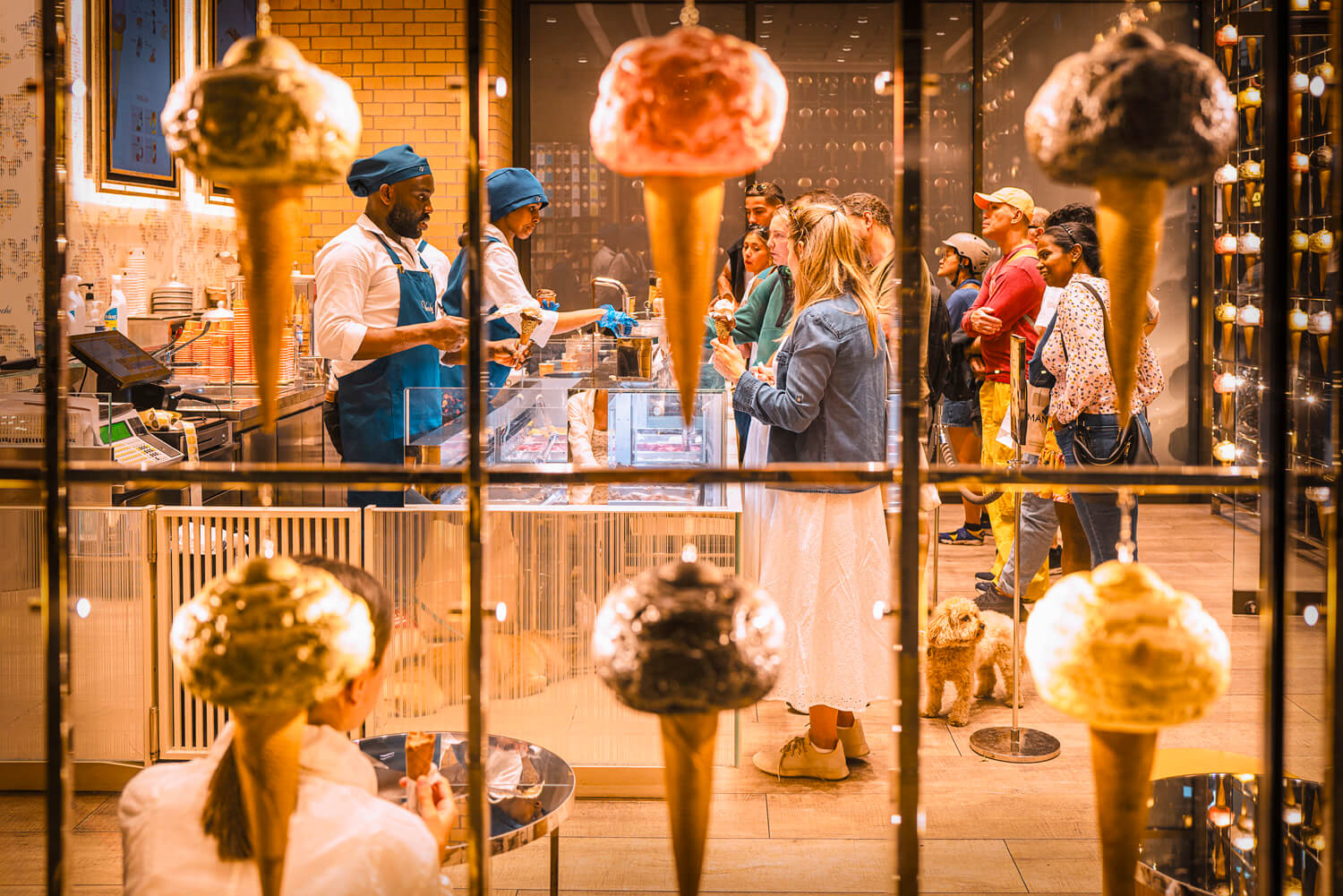 The photo presents a vibrant ice cream parlor, bustling with activity. Through the glass window, adorned with displays of ice cream cones, the interior scene unfolds with staff in blue aprons serving eager customers.