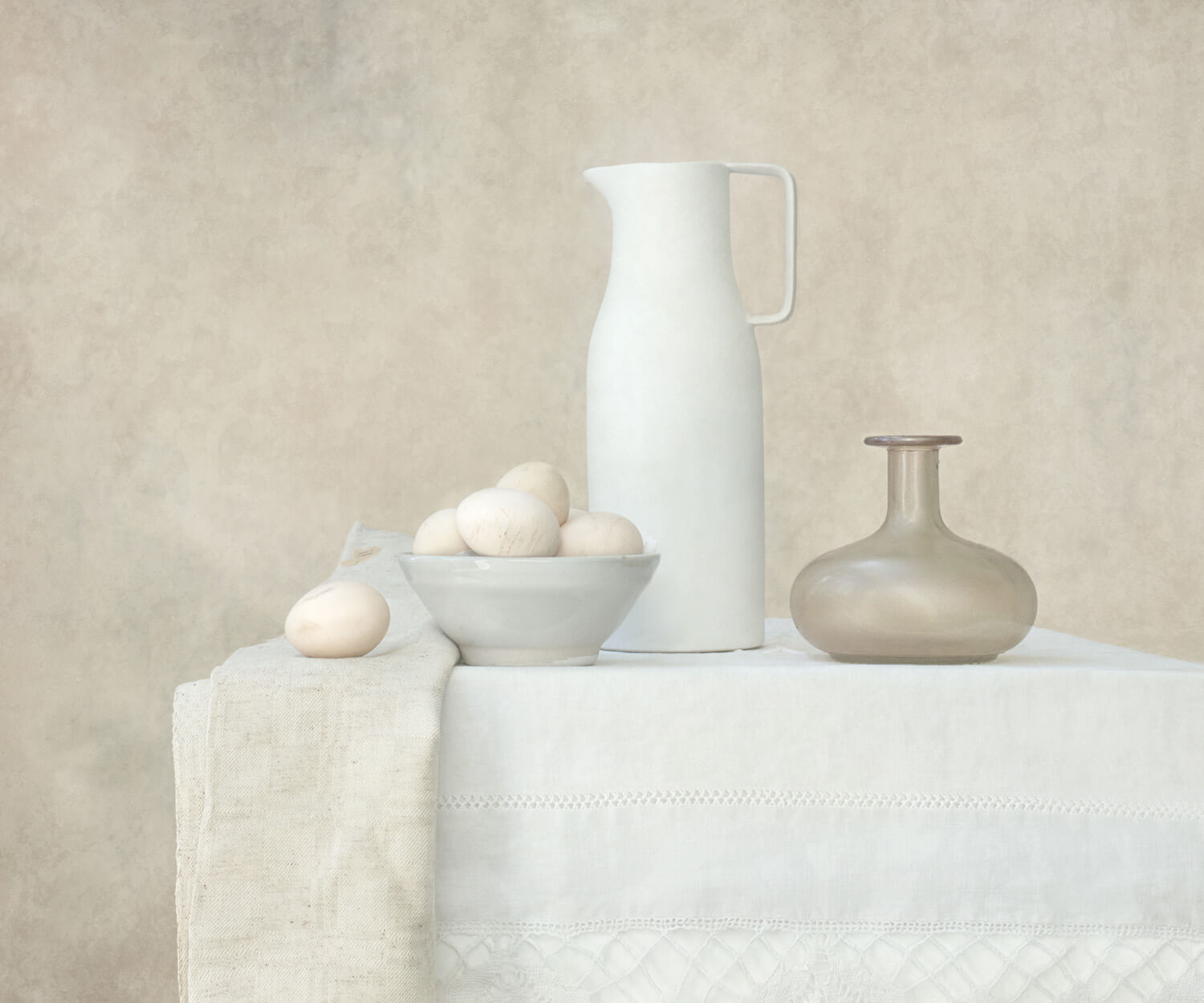 A serene still life photograph depicts a white ceramic pitcher, a bowl filled with eggs, and a small brown vase arranged thoughtfully on a table covered with a white lace-edged tablecloth. The textured background in neutral tones adds a warm, vintage ambiance to the scene, emphasizing the simplicity and beauty of the objects.