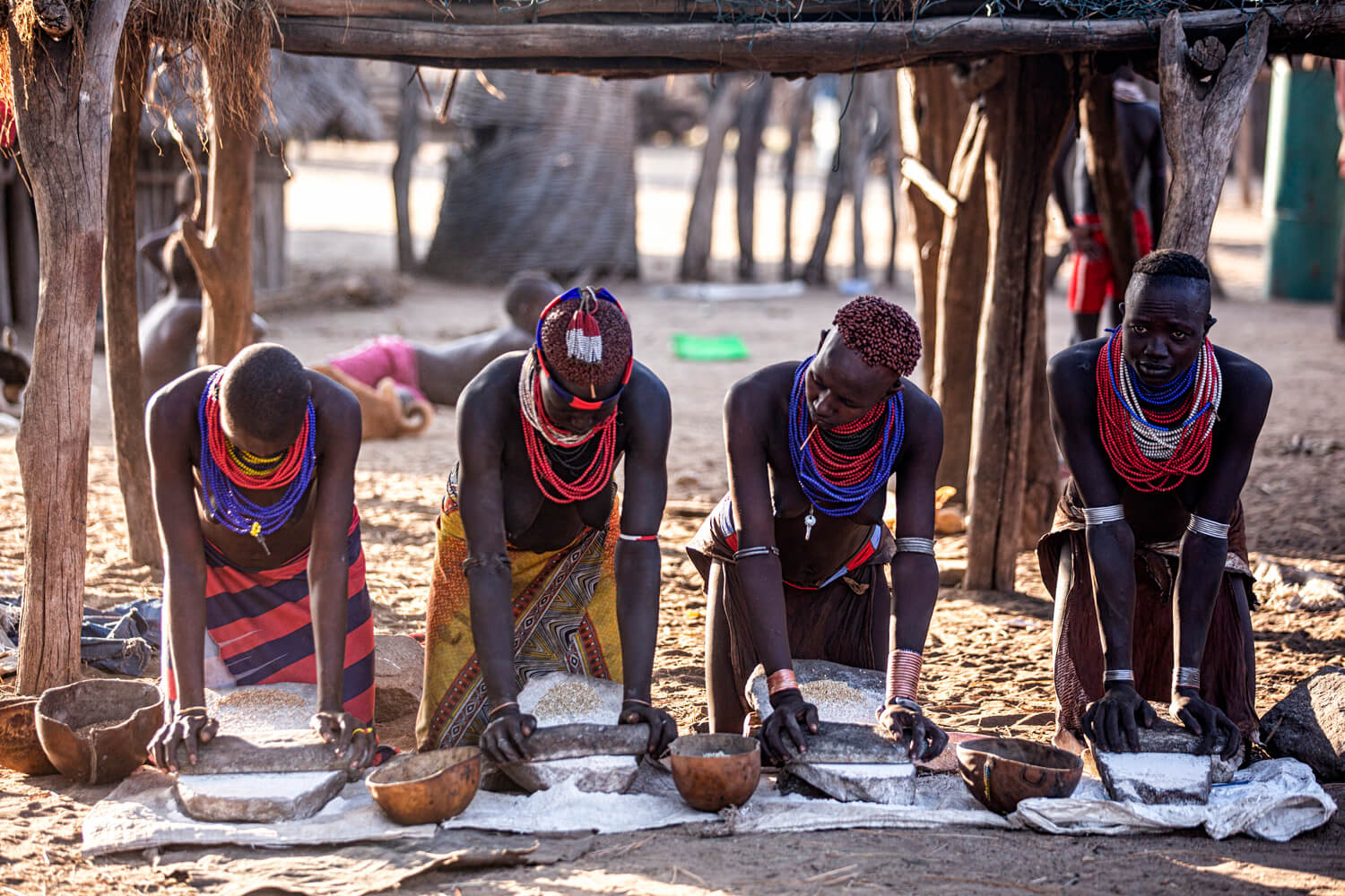 Four individuals in traditional African attire, adorned with colourful beaded necklaces, are grinding grain using large stone tools in a village setting.