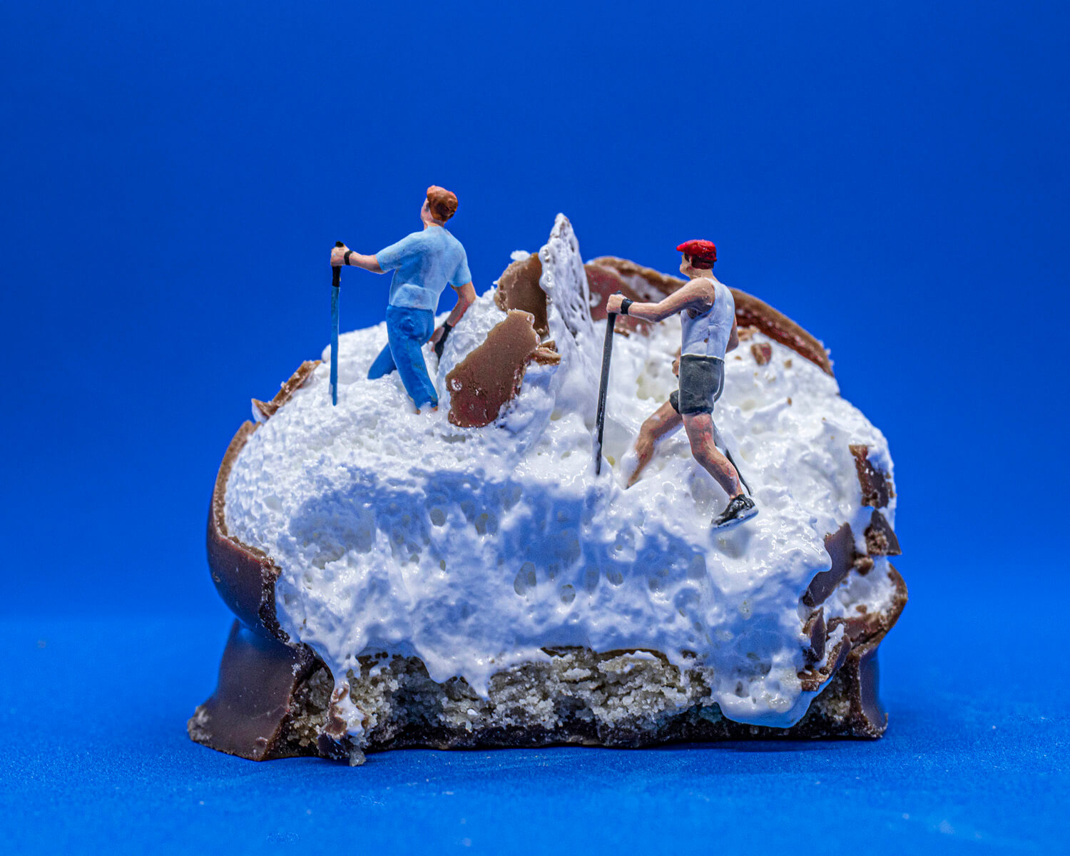 Miniature figurines of hikers traversing a rugged, snowy terrain that is actually a half-eaten tea cake, with the white marshmallow mimicking snow and the chocolatey base resembling rocky ground, against a vibrant blue backdrop.