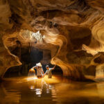 Two individuals stand in a shallow, water-filled cave, illuminated by a warm light that highlights the swirling patterns of the rock formation around them. One person holds a torch, causing sparks to reflect on the water's surface, adding a magical quality to the subterranean scene.
