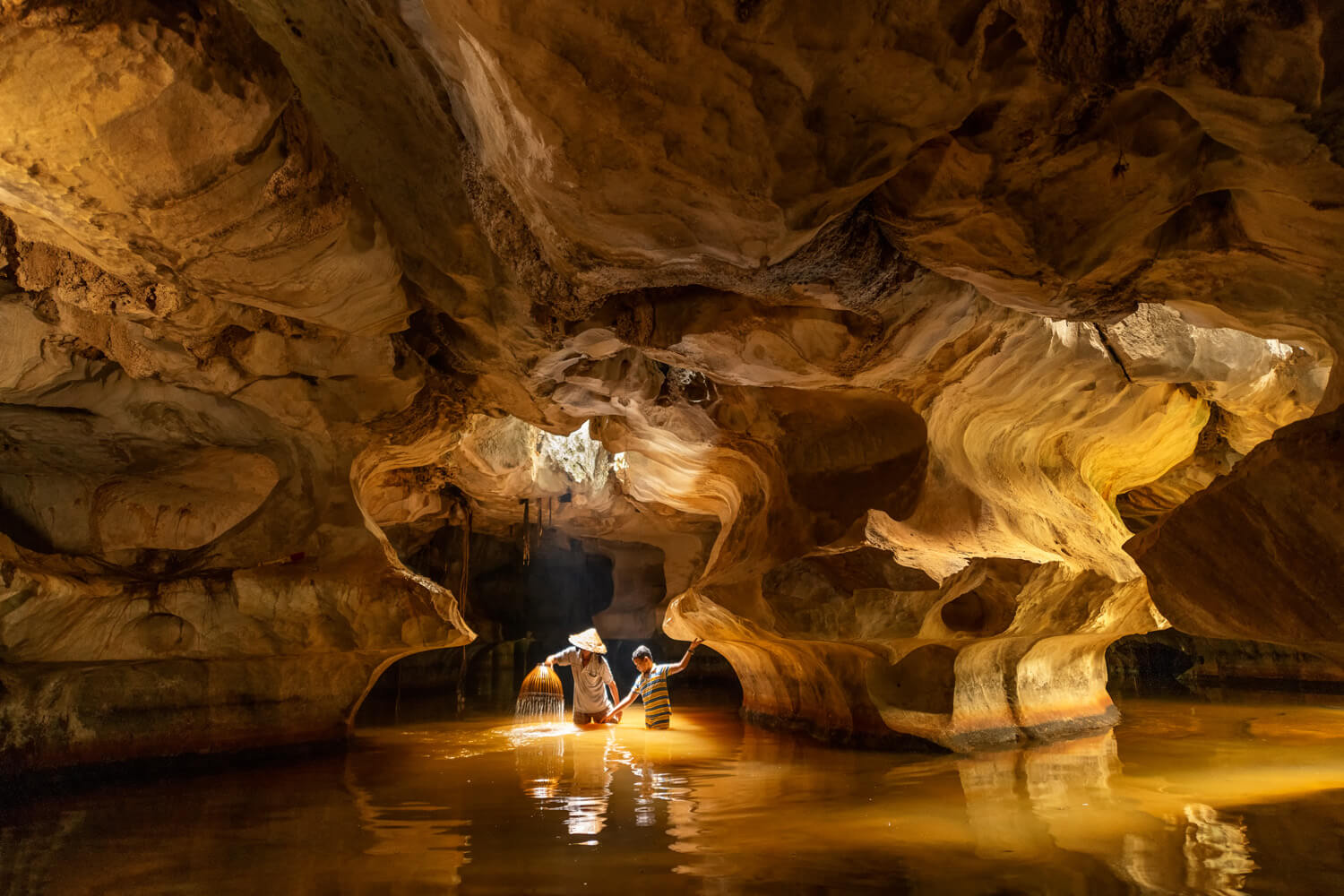 Two individuals stand in a shallow, water-filled cave, illuminated by a warm light that highlights the swirling patterns of the rock formation around them. One person holds a torch, causing sparks to reflect on the water's surface, adding a magical quality to the subterranean scene.