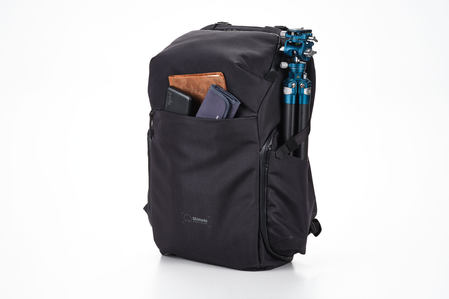 A black Shimoda Urban Explore backpack positioned upright with a tripod secured on the side and a wallet and phone protruding from an unzipped side pocket, set against a white background.