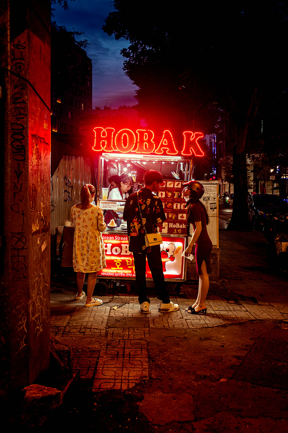 Customers gather around a brightly lit street food stall at night, the word 