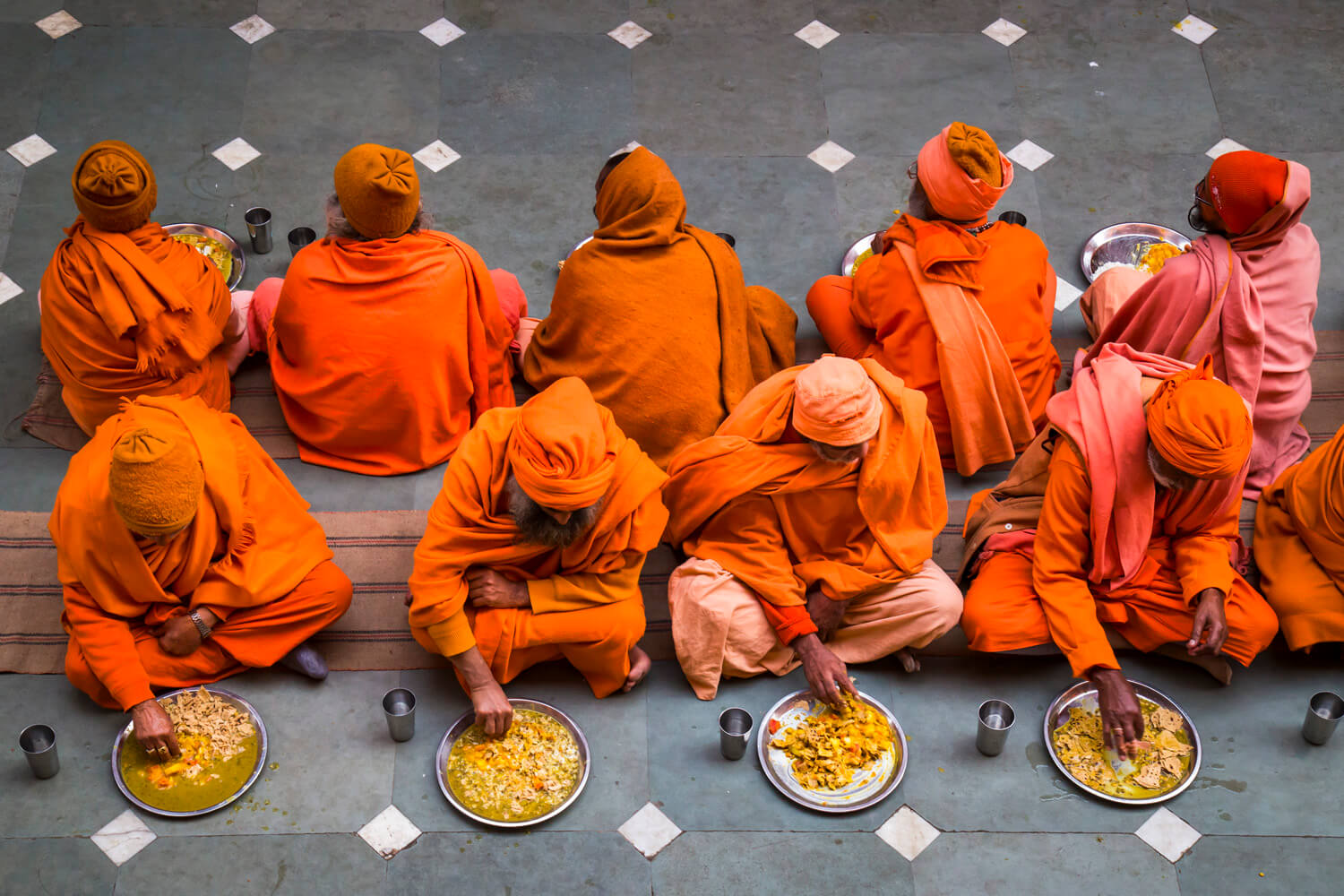A group of monks clad in vibrant orange robes are seated on the ground in a row, eating from large metal bowls. The scene is captured from above, with the monks' matching attire creating a striking contrast against the grey floor marked with white diamonds.