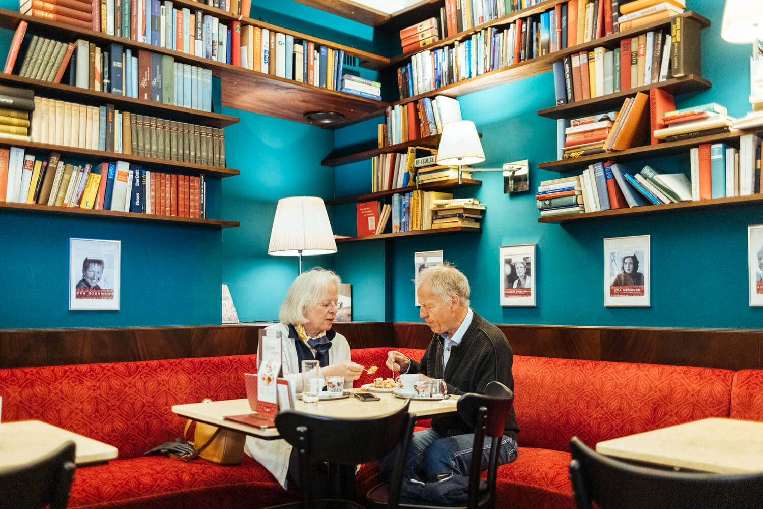 An elderly couple enjoys a meal together in a cosy café corner lined with bookshelves filled with colourful books. The vibrant teal walls are decorated with framed portraits, and the warm lighting complements the intimate ambiance.