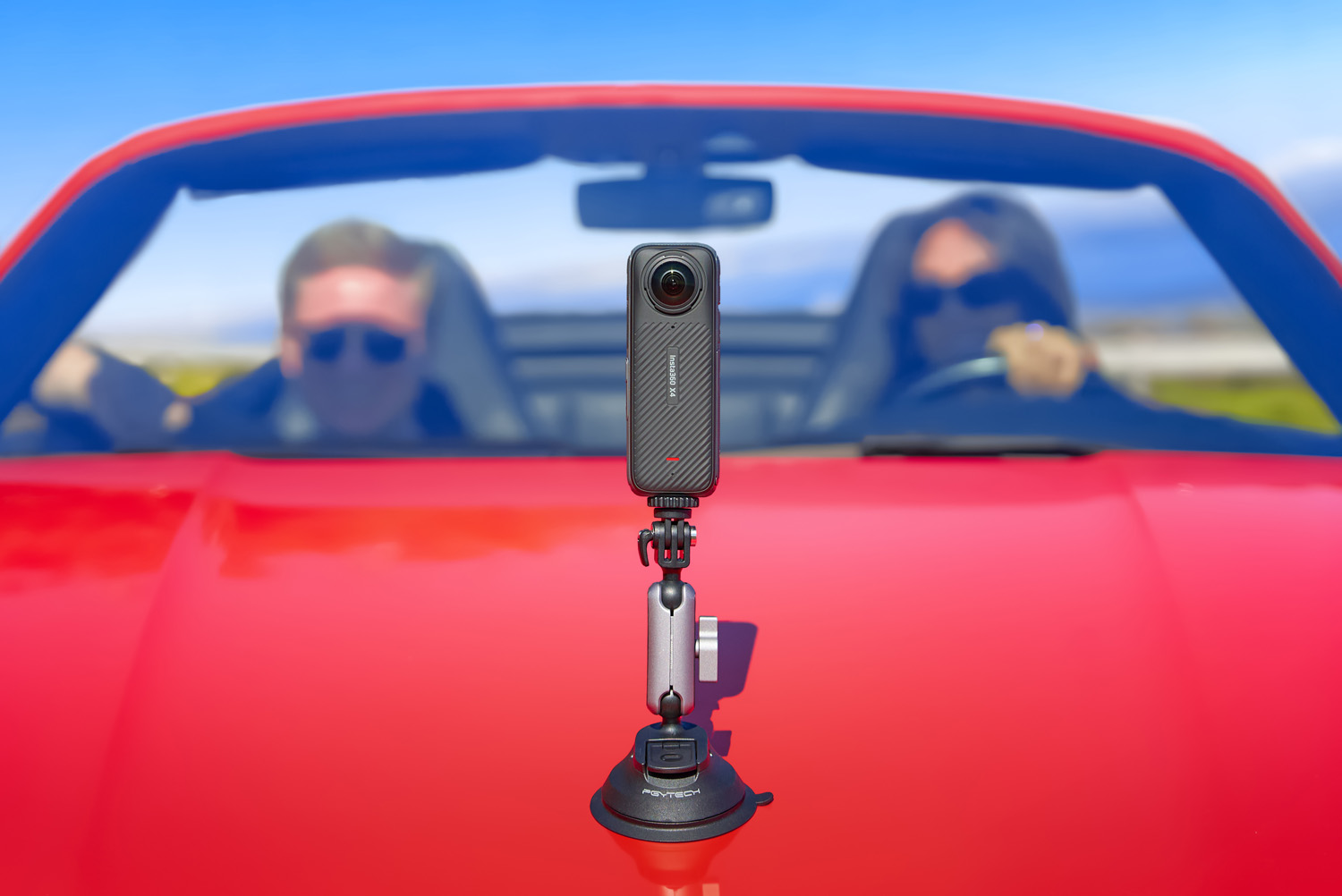 A 360-degree camera mounted on a red car's hood, with two blurred individuals in the background, one driving and the other in the passenger seat, both wearing sunglasses.
