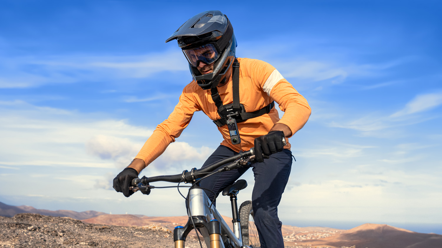 A mountain biker in an orange jersey and full-face helmet, riding on a rugged mountain trail with a 360-degree camera attached to his chest harness.