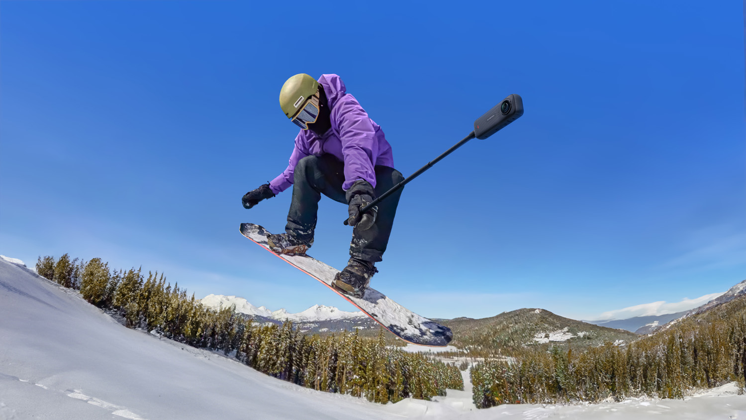 A snowboarder in mid-air during a jump, wearing a purple jacket and snow pants, with a helmet and goggles, and a 360-degree camera attached to the end of a selfie stick, with snowy mountains in the background.