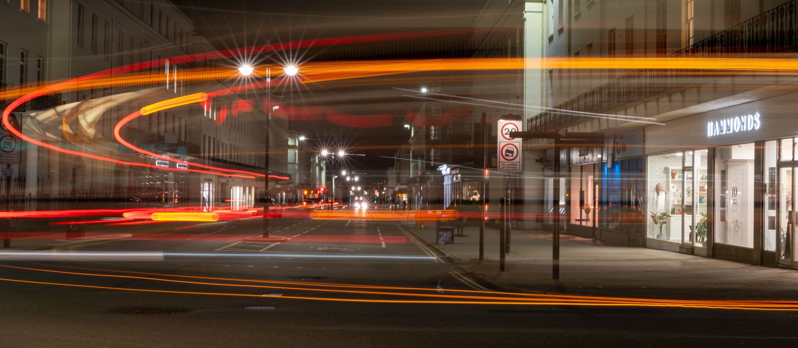 A long-exposure night shot captures the vibrant streaks of light from moving vehicles. The light trails curve through the frame, with city street scenery in the background including illuminated buildings and signage.