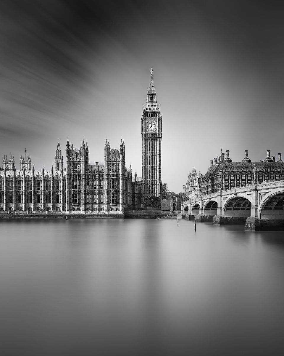 A black and white image of the iconic Big Ben and the Houses of Parliament in London. The long exposure creates a smooth surface on the River Thames and a sky streaked with motion-blurred clouds, giving the scene a timeless, grand quality.