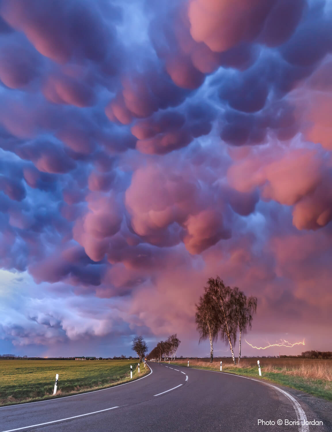 Dramatic clouds in hues of pink and purple fill the sky above a winding country road at dusk, with trees lining one side and a visible lightning strike in the distant horizon.