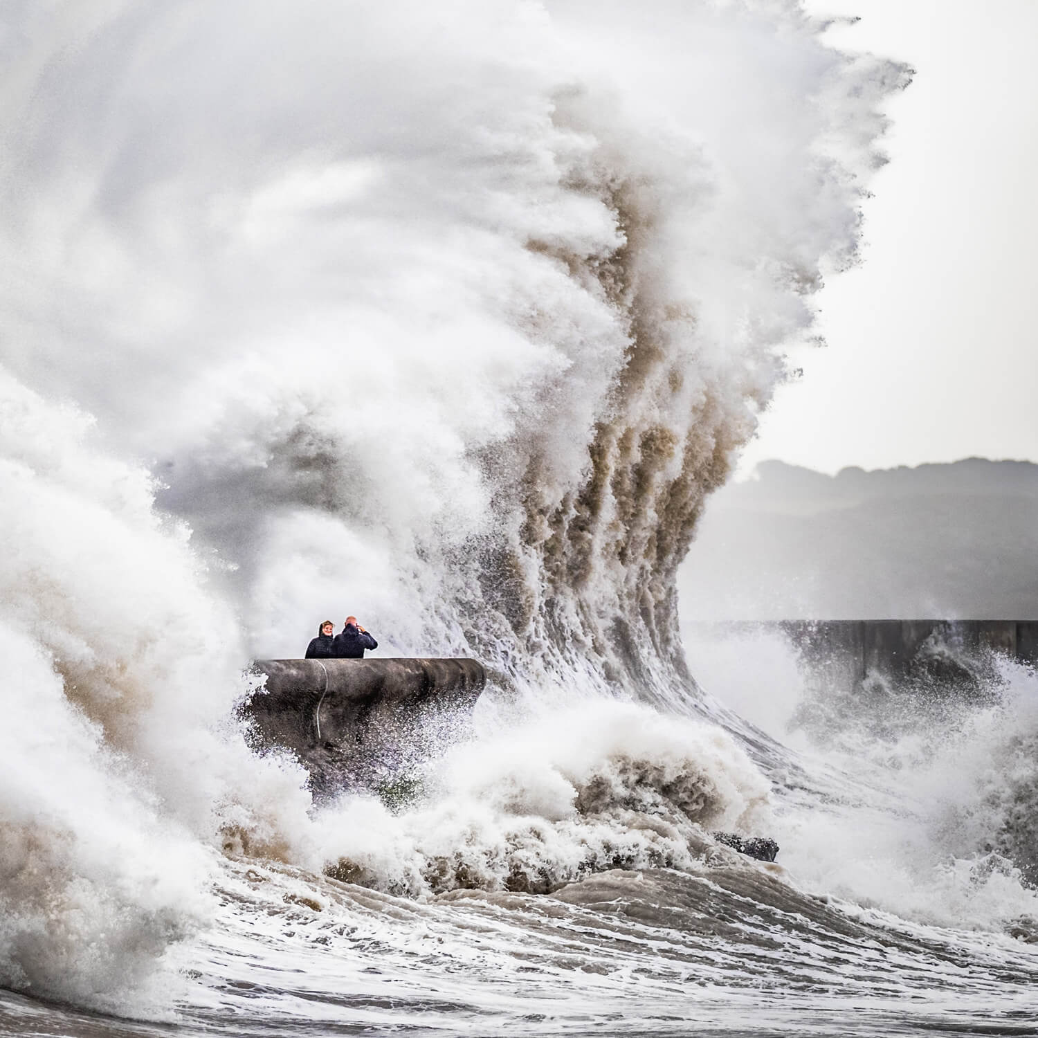 Two people standing on a concrete sea wall witness a colossal wave, resembling a liquid mountain, as it crashes over them, encapsulating the raw power of nature’s elements