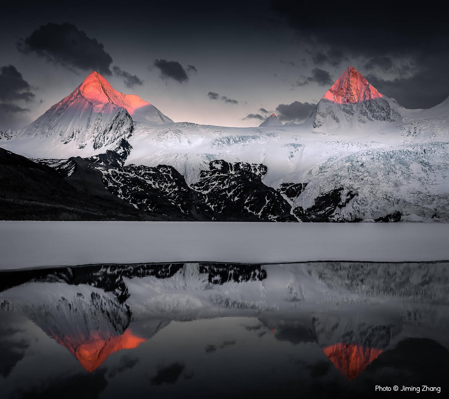 Two snow-covered mountain peaks bathed in the warm glow of sunset, with their orange-hued reflections mirrored in the calm waters of a lake below.