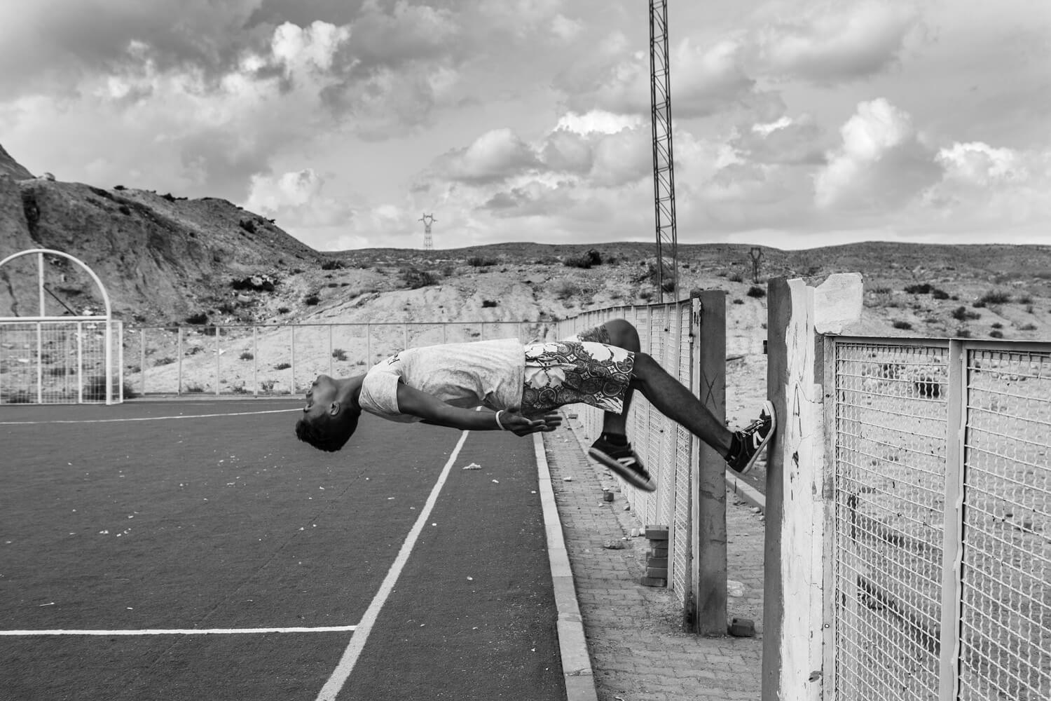 A person is frozen in time while performing a backflip off a fencepost making him look like he is performing a horizontal, blending athleticism with the stark landscape of a deserted playground.