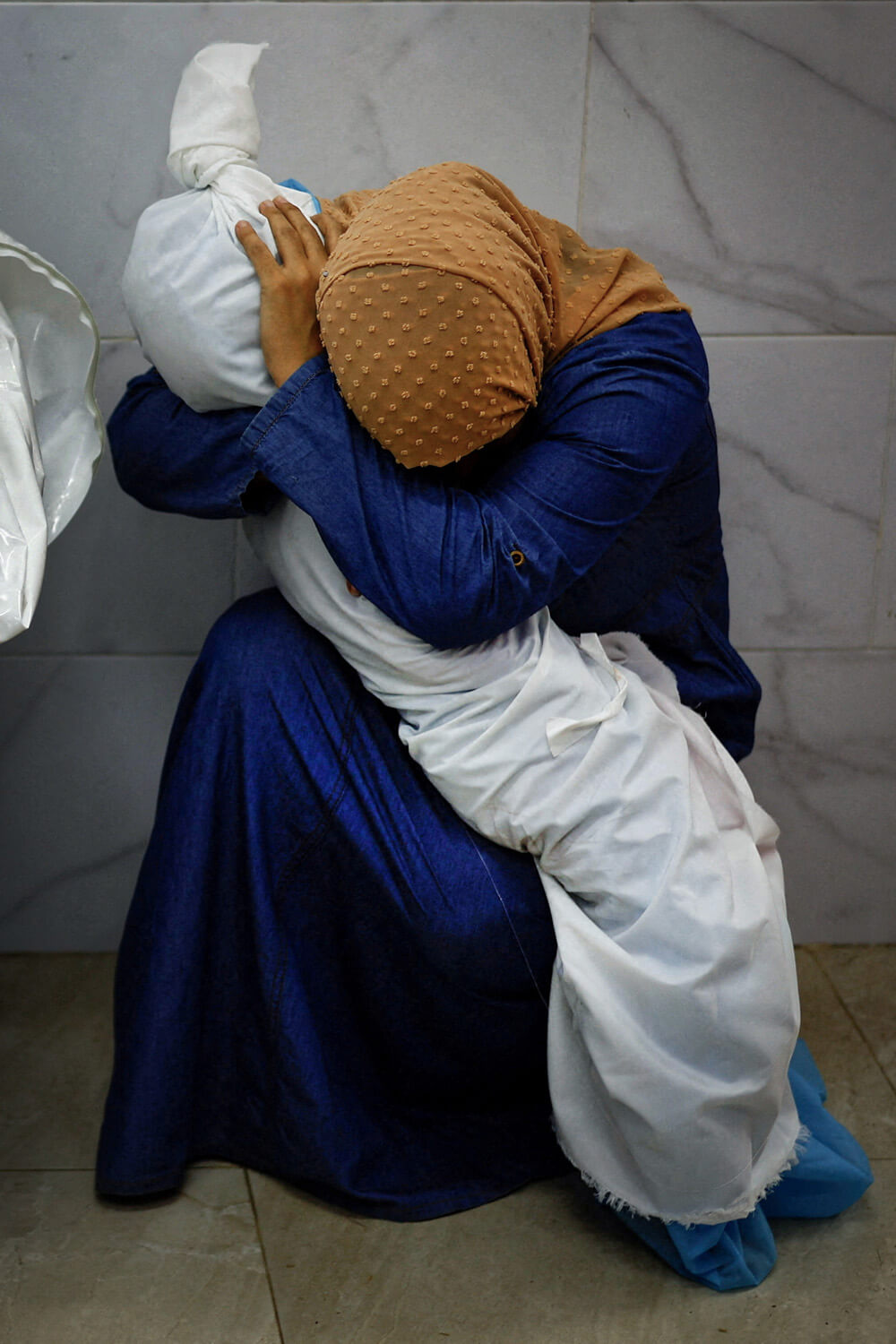 A person in a headscarf and blue garment crouches, covering their head in a posture of despair, while clutching a child's body wrapped in a white sheet