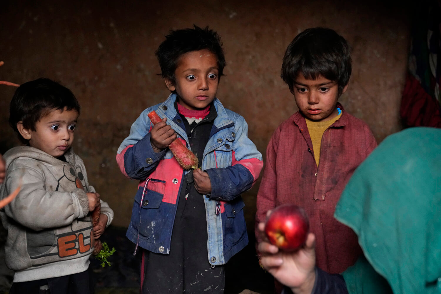 Three young children stare at a shiny red apple with expressions of bewilderment and curiosity with a dimly lit, impoverished backdrop.