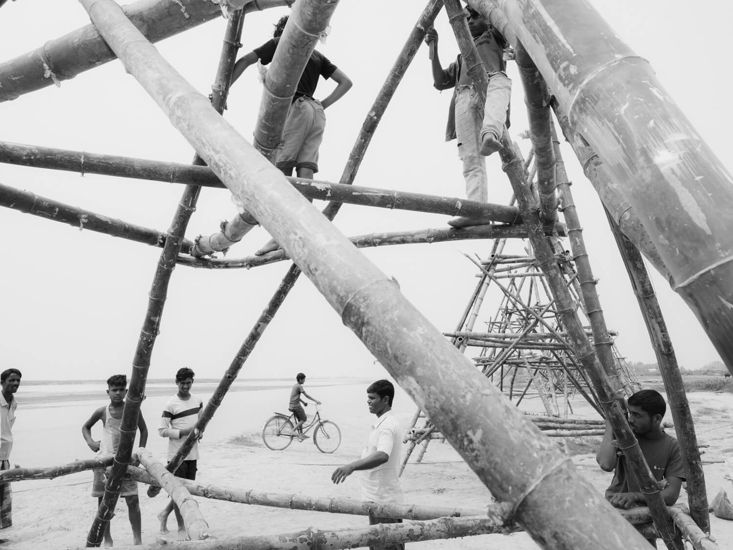 Men congregate socially on and around a large bamboo structure on a beach
