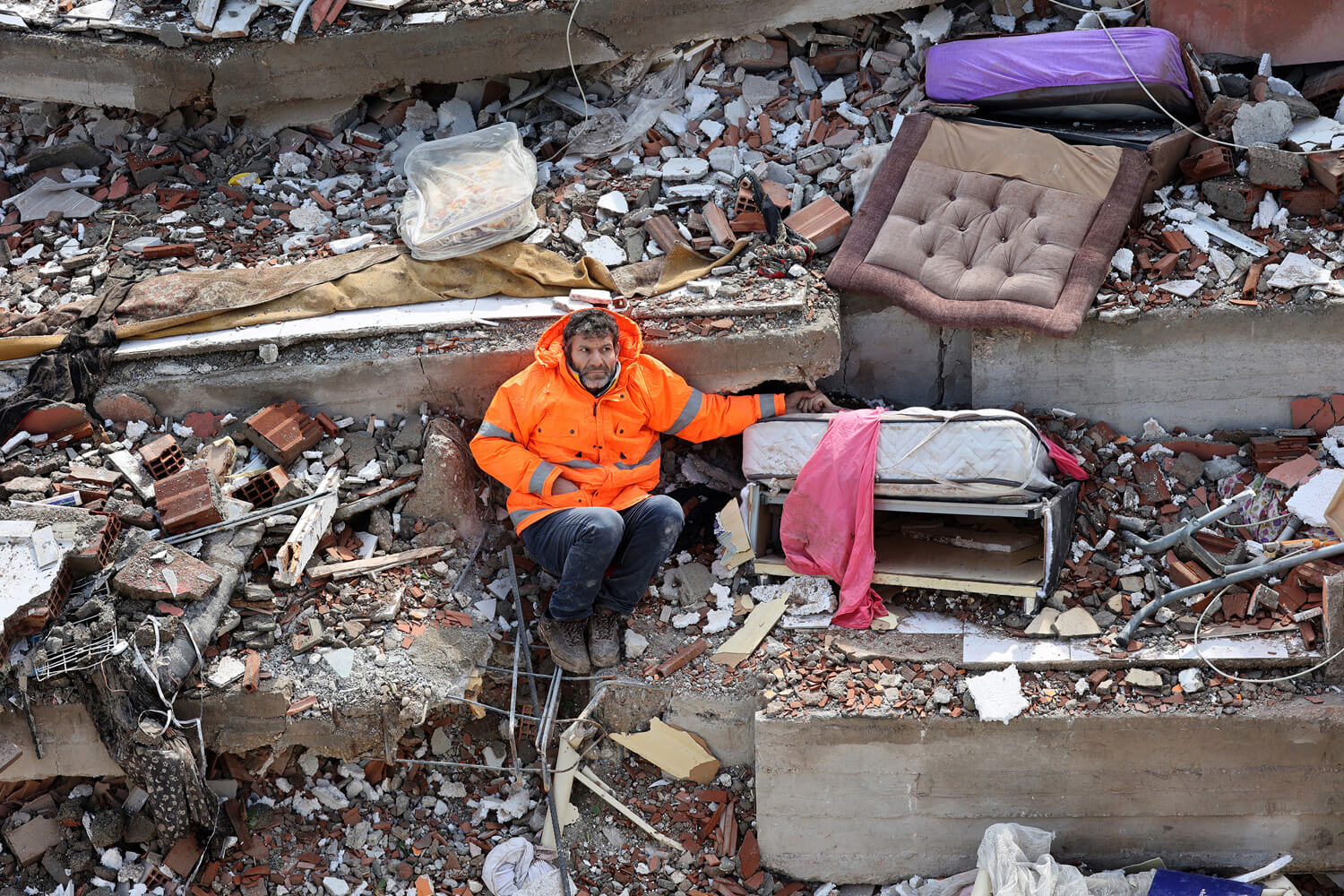 A man in a bright orange high-vis jacket sits amidst rubble, holding the hand that is protruding from beneath the concrete