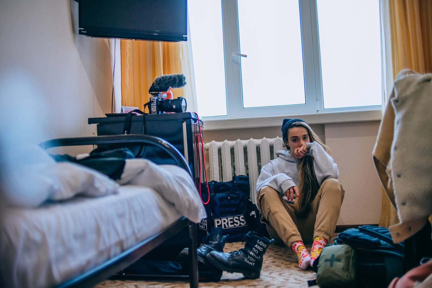 A young woman journalist sits thoughtfully on the floor, surrounded by her equipment, in a hotel room, capturing a moment of repose amidst the chaos of reporting.