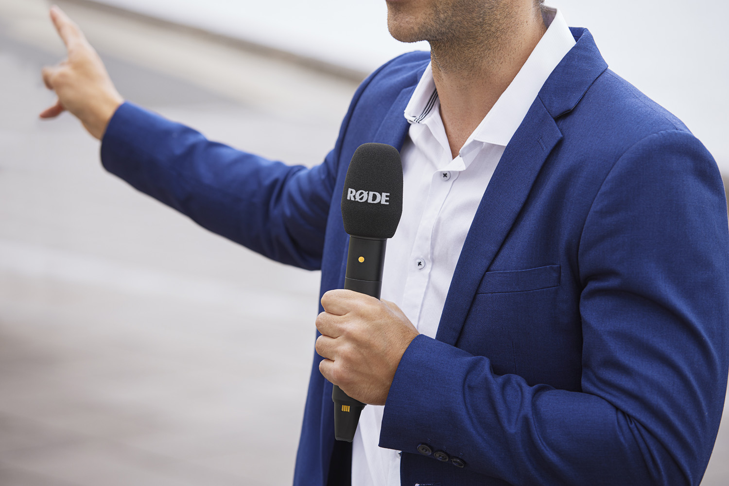 A male reporter in a blue blazer holds a black RØDE Interview PRO wireless handheld microphone. He's pointing with his right hand, directing attention off-camera, likely at an interviewee or subject of interest