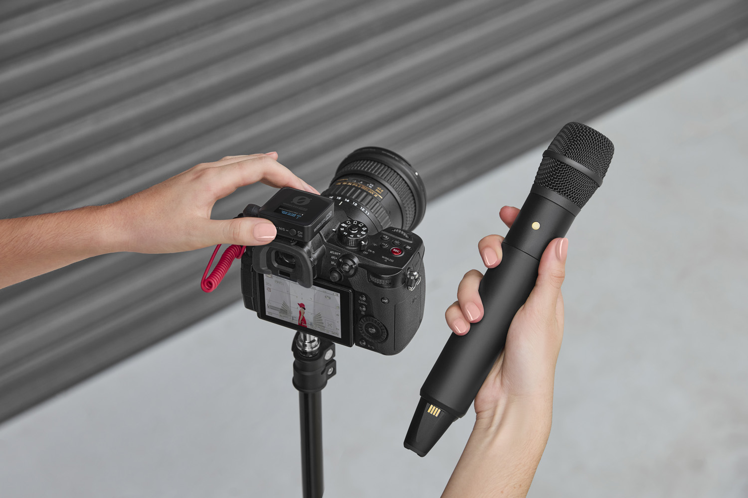 Close-up of a hand holding the RØDE Interview PRO wireless handheld microphone near a DSLR camera mounted on a tripod, highlighting the pairing process between the microphone and the camera