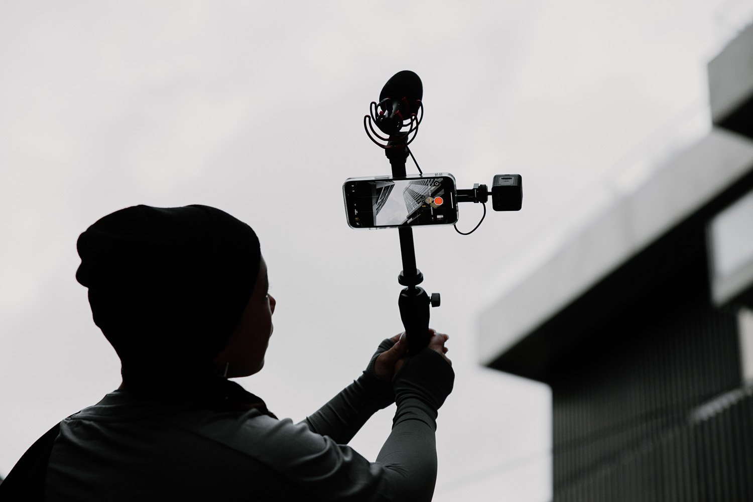 Silhouette of a person wearing a beanie, holding up a mobile filmmaking setup against a cloudy sky. The setup includes a smartphone mounted on a handheld stick mount, equipped with a RØDE microphone and an external light source. The backdrop suggests an urban setting, ideal for on-the-go content creation