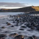 A serene landscape photograph capturing a tranquil beach scene at dusk with smooth, long exposure waters flowing over rounded pebbles. In the background, the ruins of Dunstanburgh Castle stand atop a grassy cliff, bathed in the warm glow of the setting sun. The sky is partly cloudy, hinting at the closing of a clear day