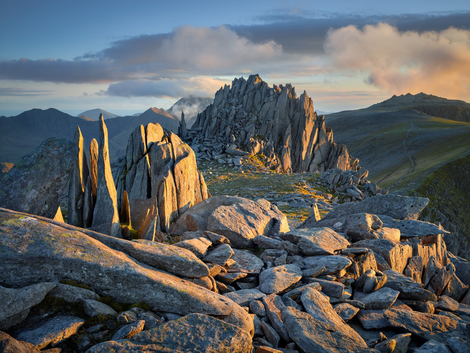 Dynamic landscape photograph showcasing rugged, jagged rock formations, known as the 'Fortress of the Four Winds,' under a soft golden hour light. The foreground is littered with large, angular boulders, while in the background, a range of pointed mountain peaks stretches under a sky with wispy clouds at sunset.