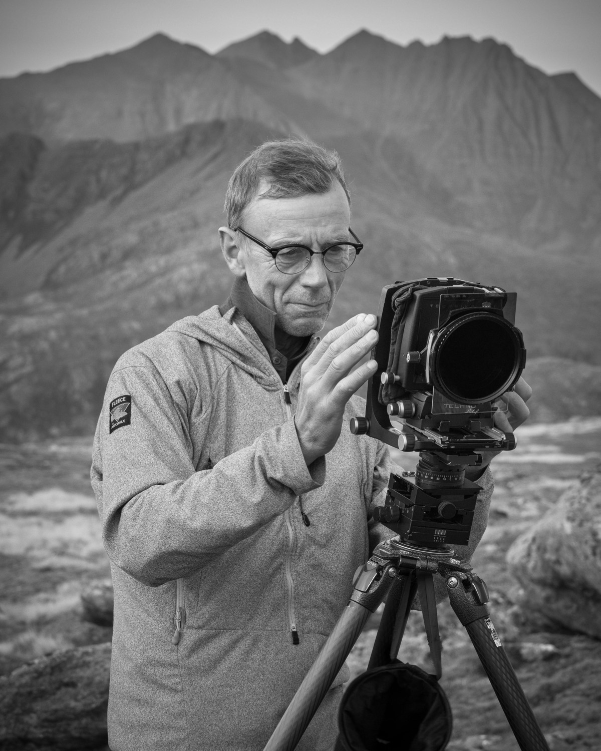 Black and white portrait of Joe Cornish, a mature, bespectacled man, thoughtfully inspecting his large format camera perched on a tripod. He stands in a rugged outdoor setting with a backdrop of dramatic mountain ranges, embodying a focused and professional demeanor.