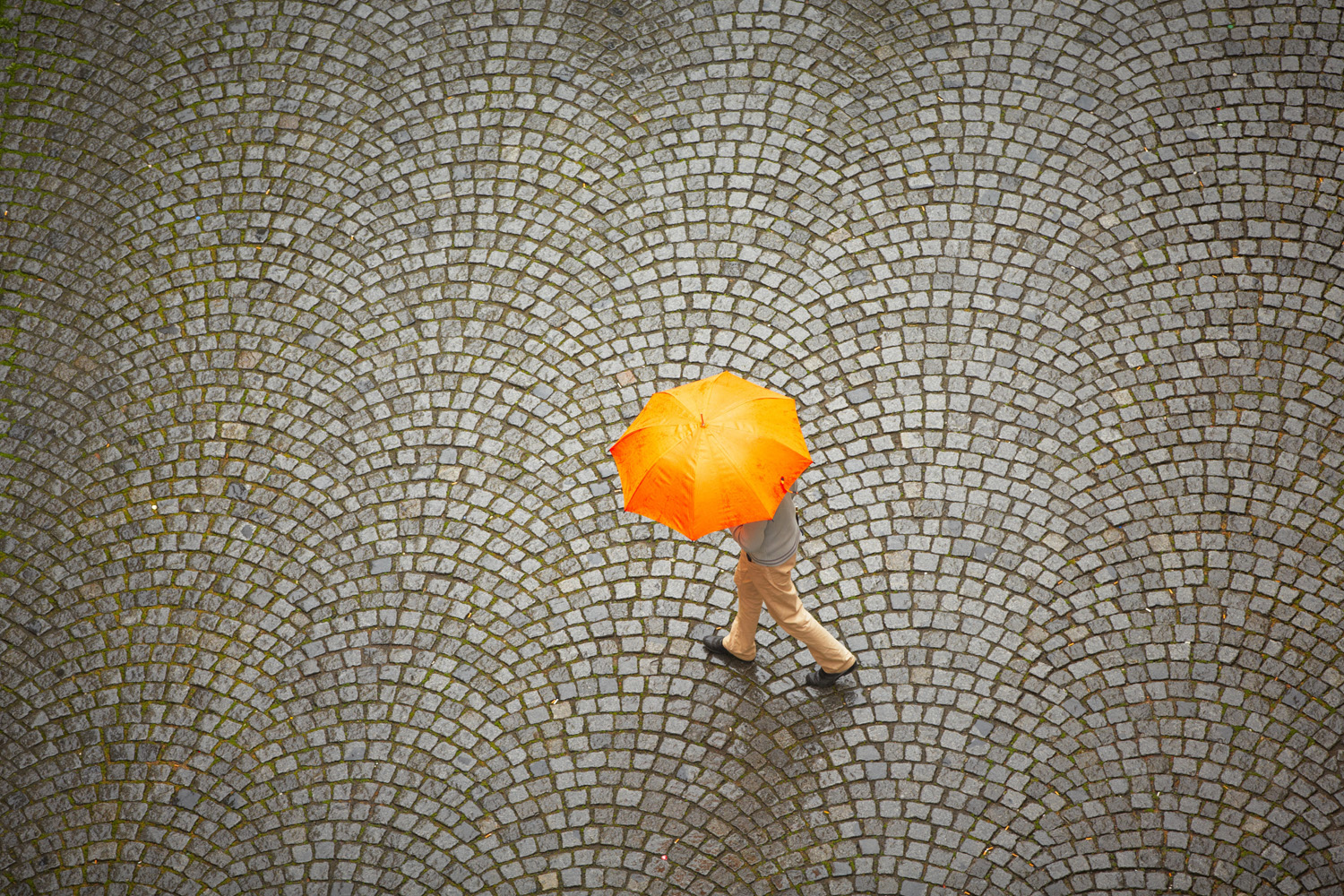 High shot of man with orange umbrella in rain against patterned stone pavement