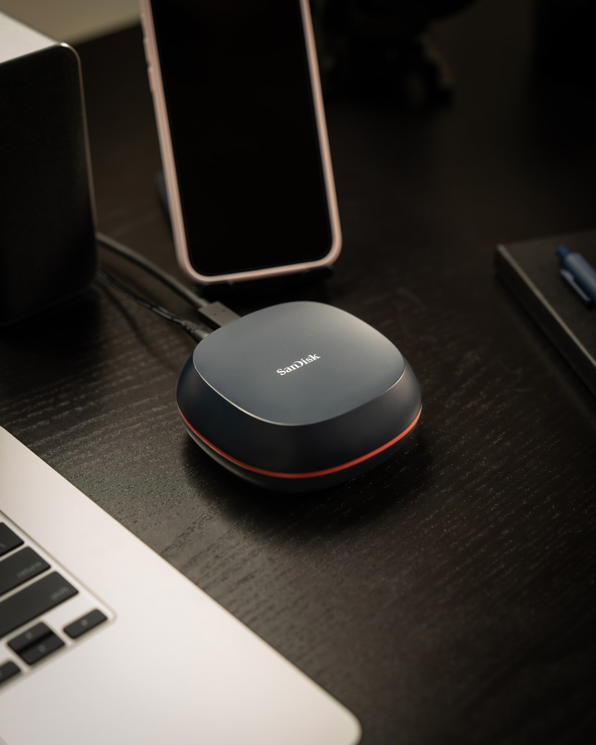 A close-up view of the SanDisk Desk Drive SSD on a dark wooden desk, connected to a laptop via a USB cable, with a smartphone propped up next to it. The drive features a sleek design with a grey top and an orange accent line.